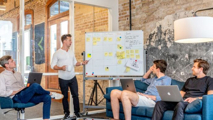 three men sitting while using laptops and watching man beside whiteboard