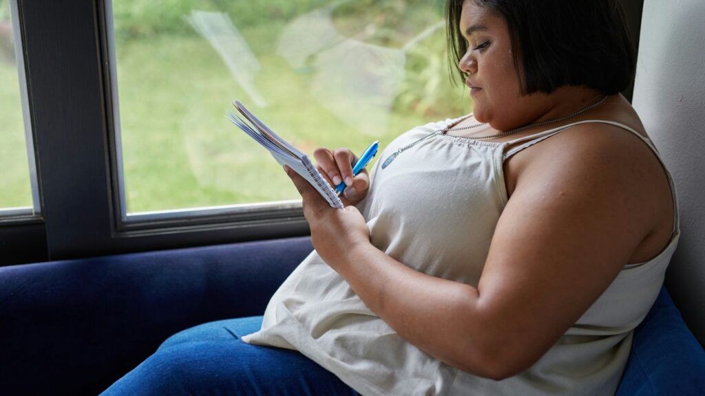 A woman writes in a notebook by a window.