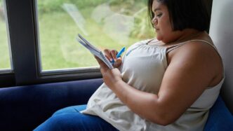 A woman writes in a notebook by a window.