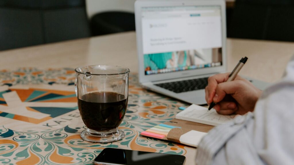 writing person beside glass filled with black liquid on turned off iPhone in front of turned on MacBook Air on table