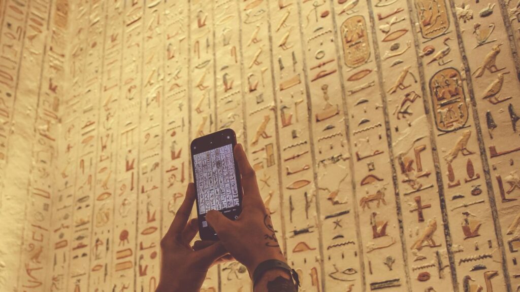 A person using a cell phone in front of a wall with egyptian writing