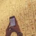 A person using a cell phone in front of a wall with egyptian writing