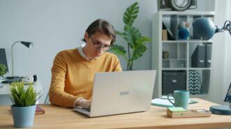 Man in yellow sweater working on laptop at desk.