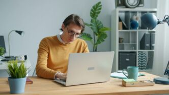 Man in yellow sweater working on laptop at desk.