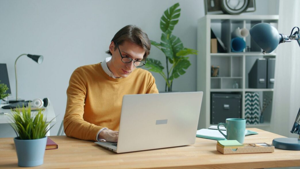 Man in yellow sweater working on laptop at desk.