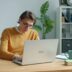 Man in yellow sweater working on laptop at desk.