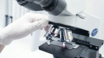 A person in white gloves looking through a microscope