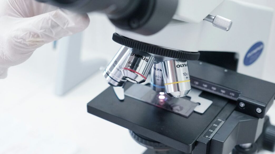 A person in white gloves is looking through a microscope