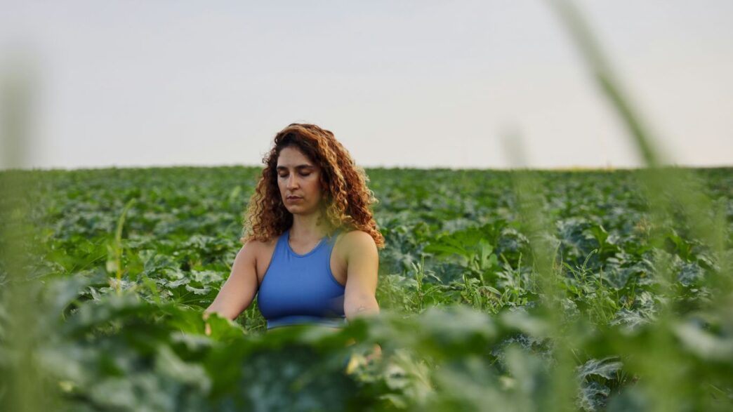 selective focus photography of woman meditating while sitting on ground surrounded by plants during daytime