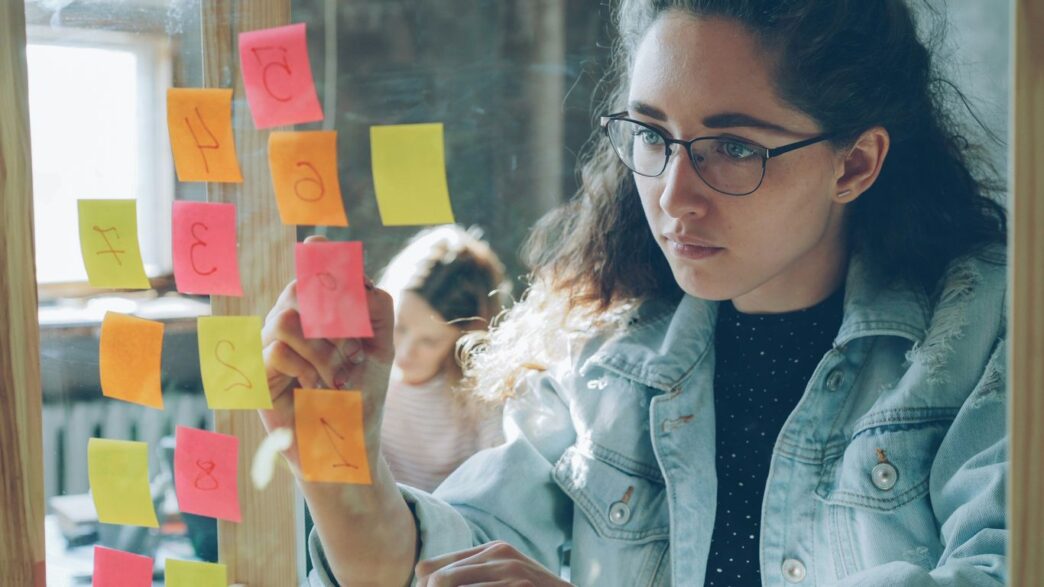 Woman analyzes colorful sticky notes on glass.