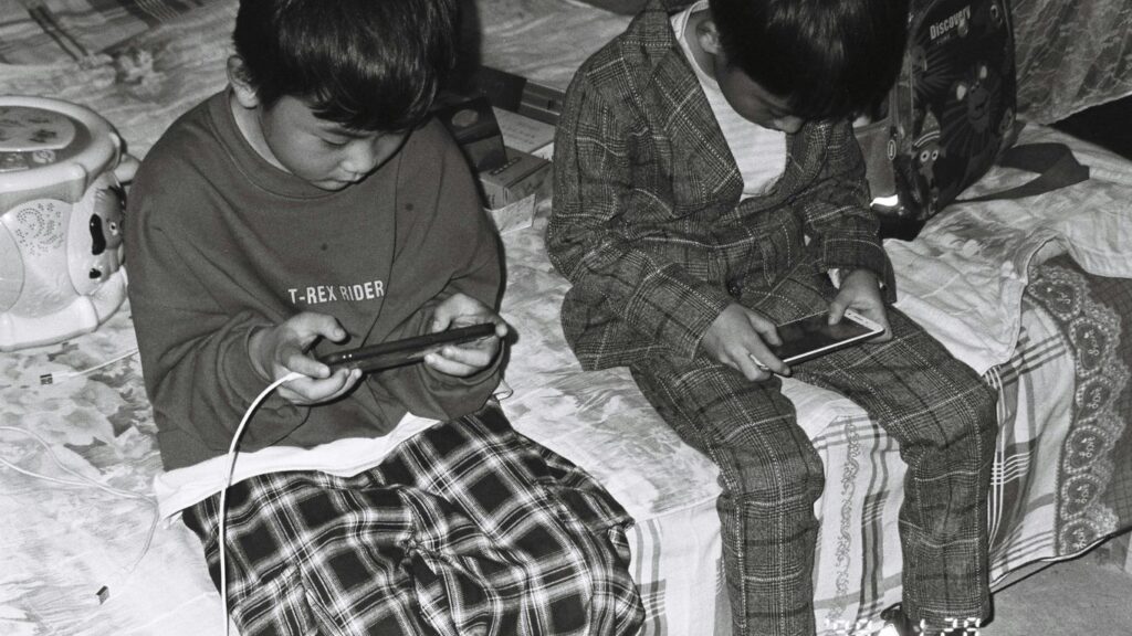 two young boys sitting on a bed looking at their cell phones