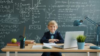 Young boy smiles at desk with laptop and chalkboard.