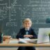 Young boy smiles at desk with laptop and chalkboard.