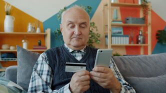 Elderly man holding credit card and smartphone for online shopping.