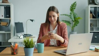 Young woman smiling while using her smartphone at desk.