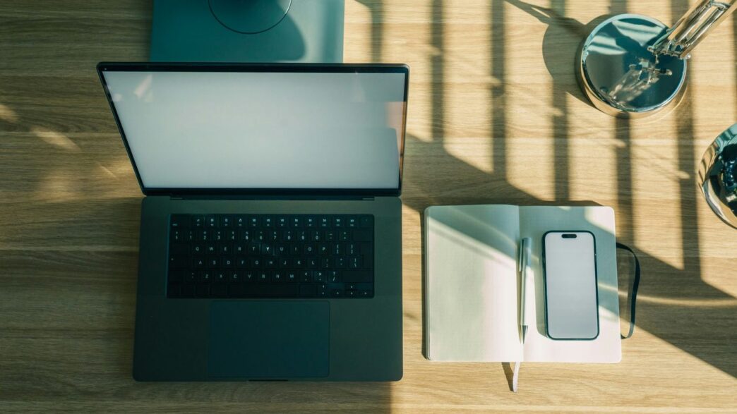 Laptop, phone, and notebook on a wooden desk.