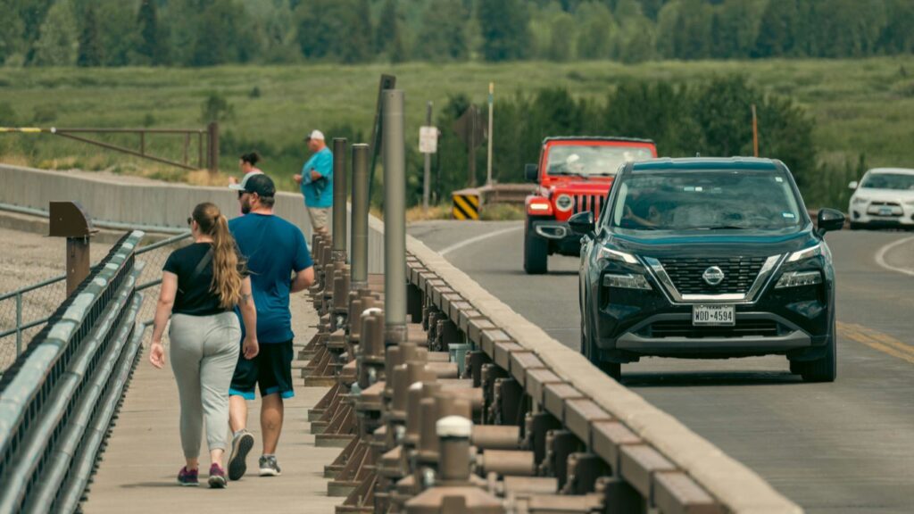 People walk on a bridge with cars driving by.
