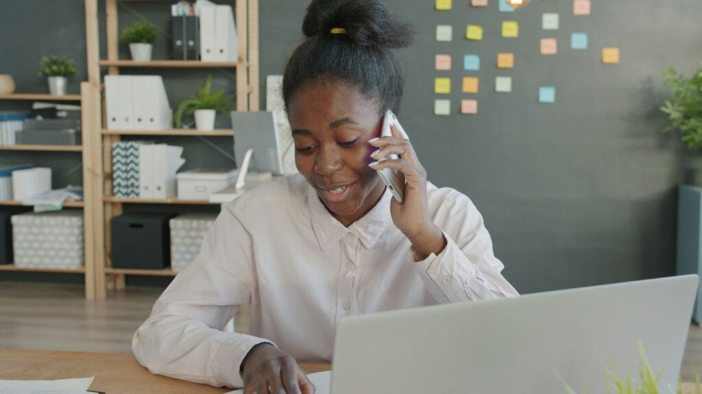 Young woman talking on phone at office desk with laptop.