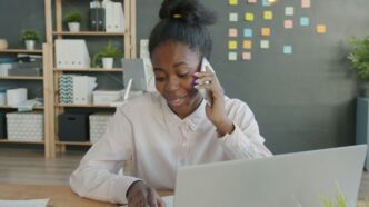 Young woman talking on phone at office desk with laptop.
