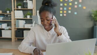 Young woman talking on phone at office desk with laptop.