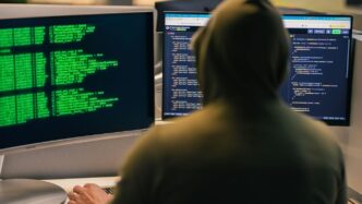 Hacker in hoodie working on multiple computer screens