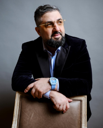 man in a black coat while wearing a blue watch and leaning on a chair