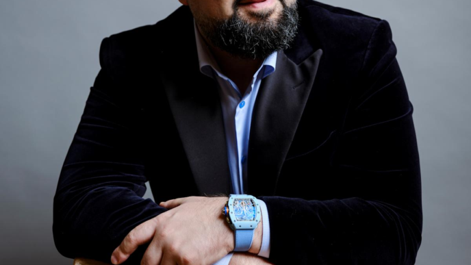 man in a black coat while wearing a blue watch and leaning on a chair